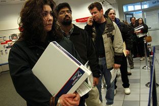 Customers waiting in line at a U.S. Post Office counter, one holding a Priority Mail box